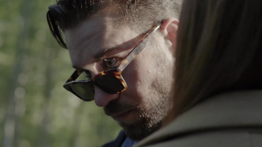 A young man wearing stylish sunglasses a trendy haircut with stubble looks intensely over the top of his sunglasses as he stands in front of a woman in a coat