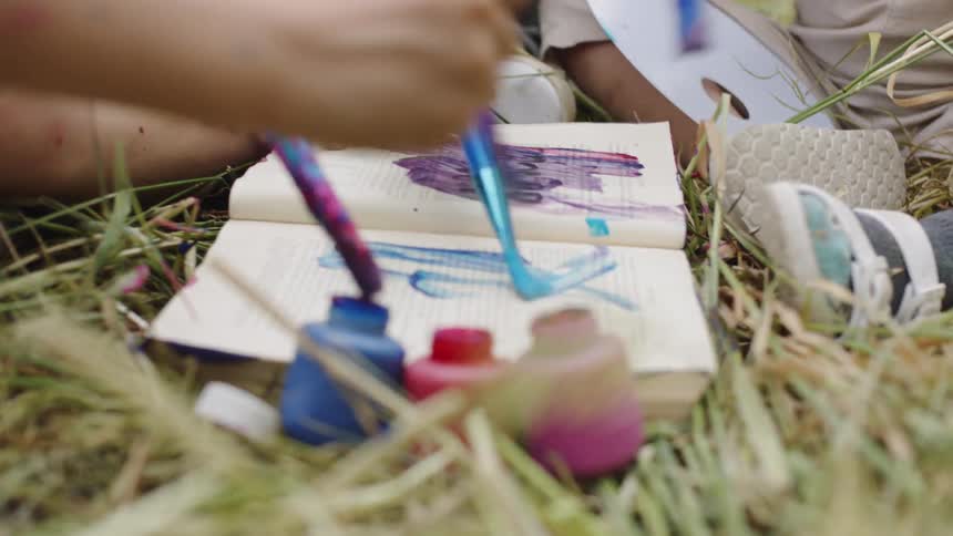 Children painting on notebook paper while sitting in the grass