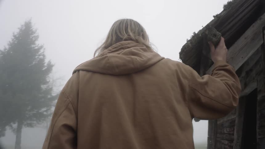 Long hair blonde man holds the roof of a hut and take a look at inside in the countryside on a foggy day