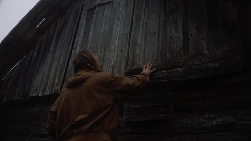 Blond man with long hair touches the wooden hut on a cloudy day