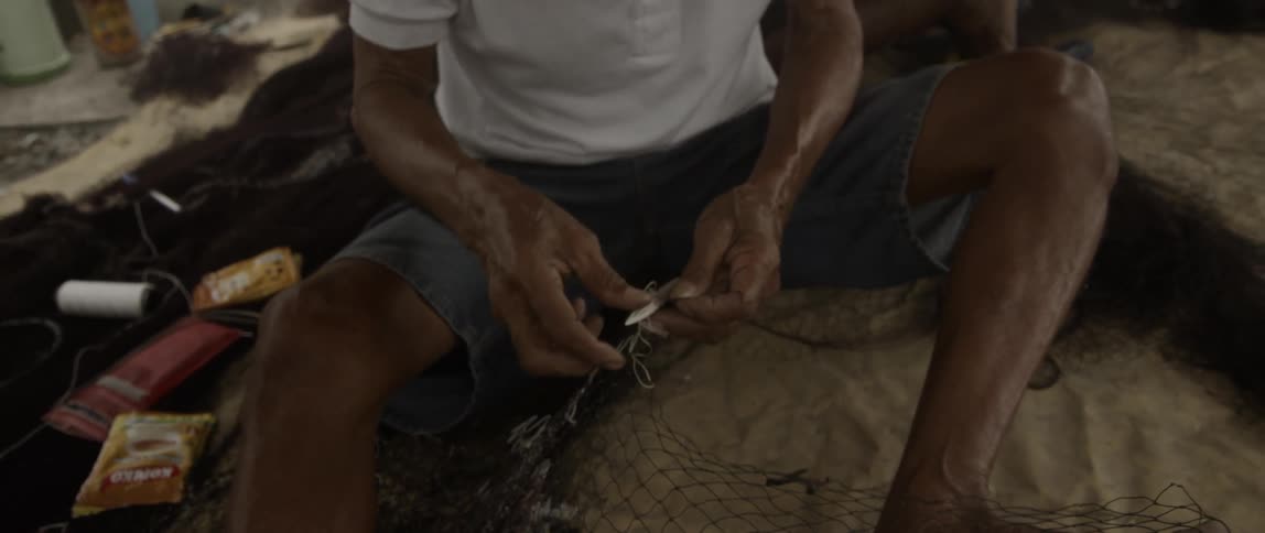 Man sitting on sand sewing together a broken fishing net