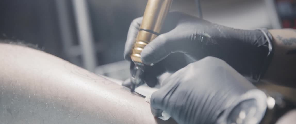 A tattooist wearing black gloves uses a tattoo gun to apply ink to a mans leg
