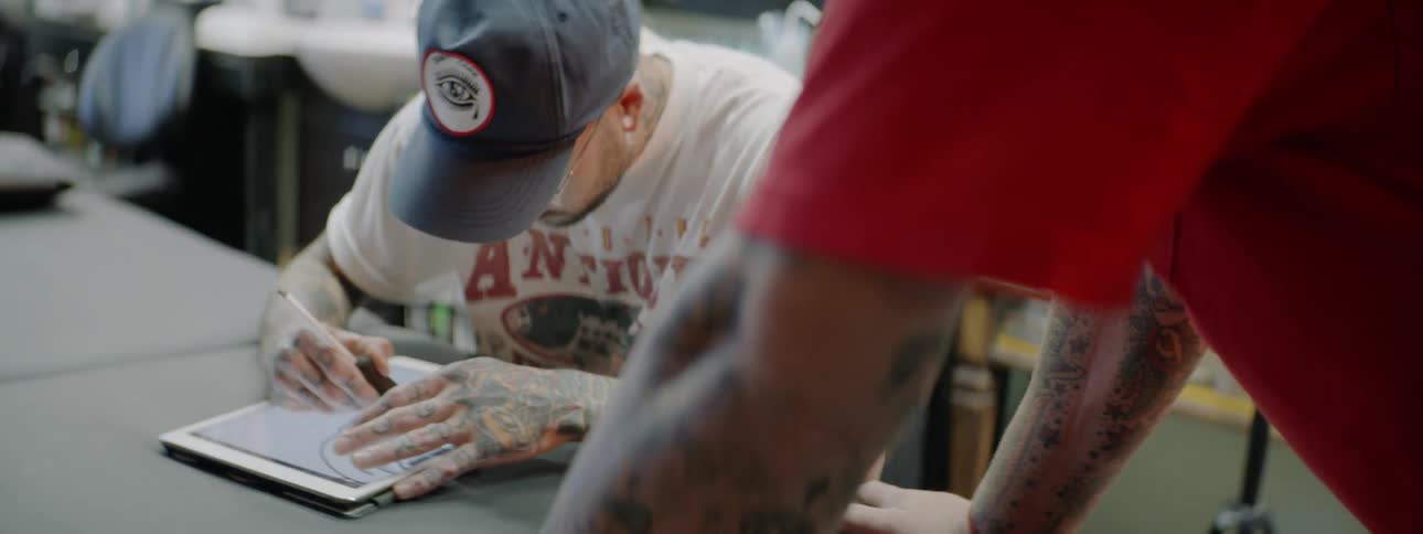 Tattooed man wearing cap draws on tablet on the table while another man stands next to him during the daytime