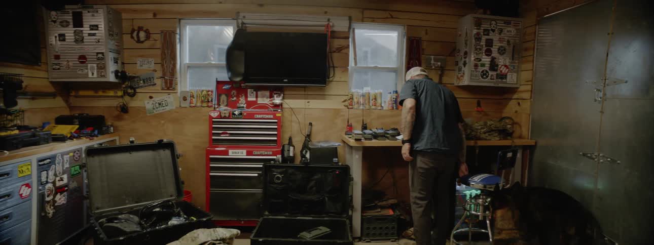 Man walks across cluttered workshop and sits down on chair while german shepherd watches