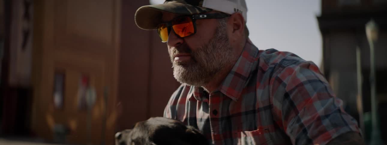 A man wearing sunglasses and a hat is petting a black dog that is licking his face