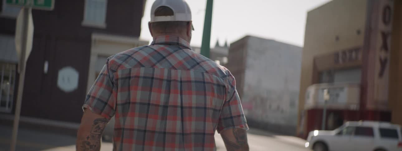 Tattooed man walks his dog on sidewalk as white automobile turns onto city street lined with old buildings in the afternoon