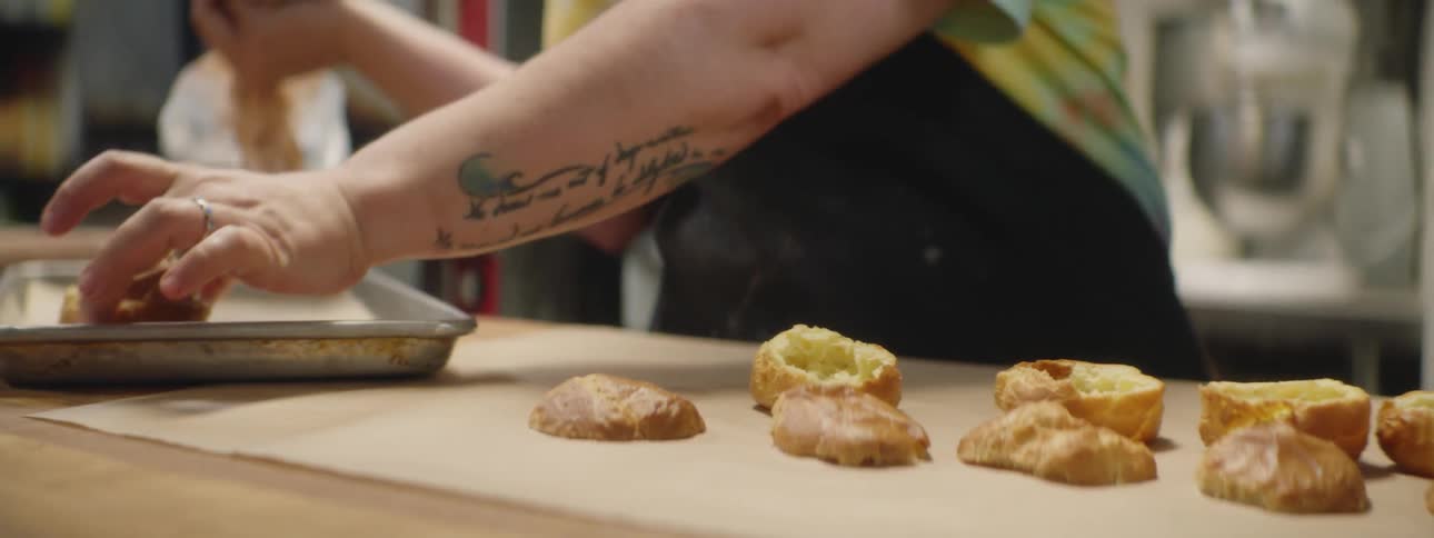 A man is carefully applying frosting to a small pastry and places it on a baking pan while more of the unfrosted pastries sit on a counter to the side