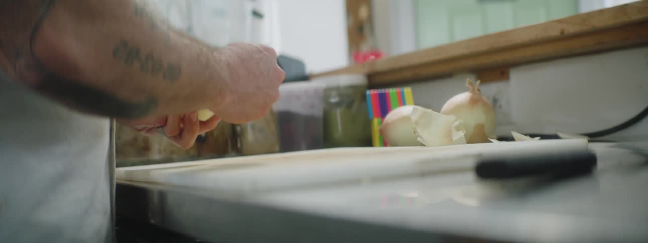 Tattooed man peels an onion in the kitchen during the daytime