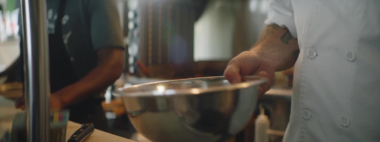 A cook with an arm tattoo pours powder into a steel bowl and swishes it around