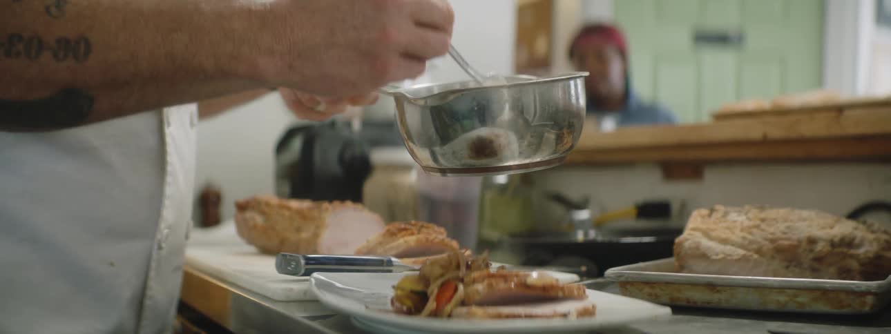A cook with an arm tattoo spreads sauce on slices of meat with a spoon