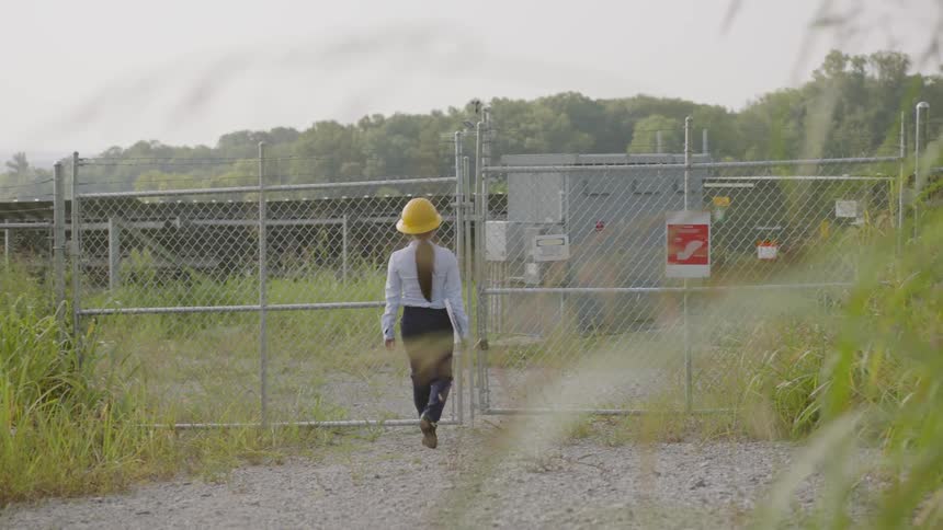 Woman wearing a safety hat walks towards a fenced in area carrying a clipboard