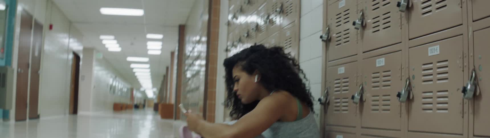 Curly hair girl with bare foot sits down on the floor in front of the school locker uses smart phone in the school hall