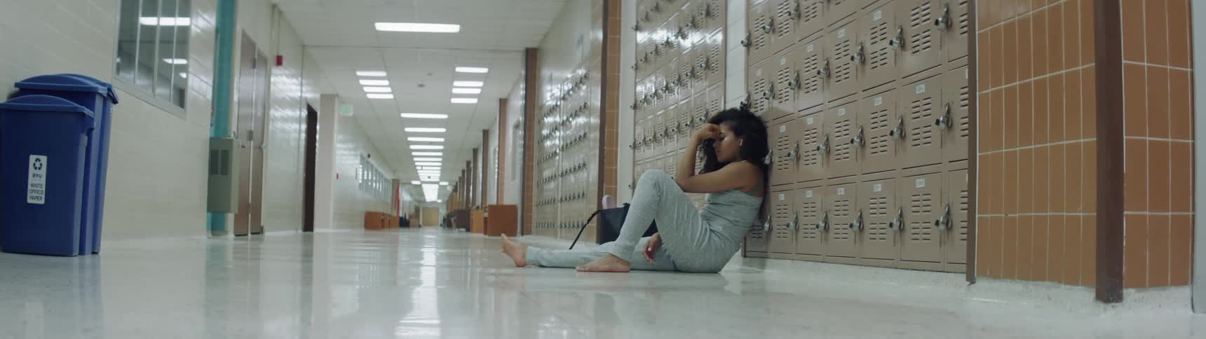 A distressed female is sitting on a linoleum floor in the school hallway by herself with her back leaning against a set of lockers
