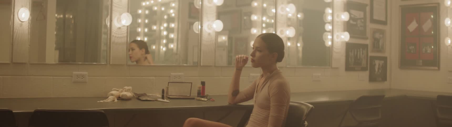 A dancer sits in front of mirrors with lights in a dressing room