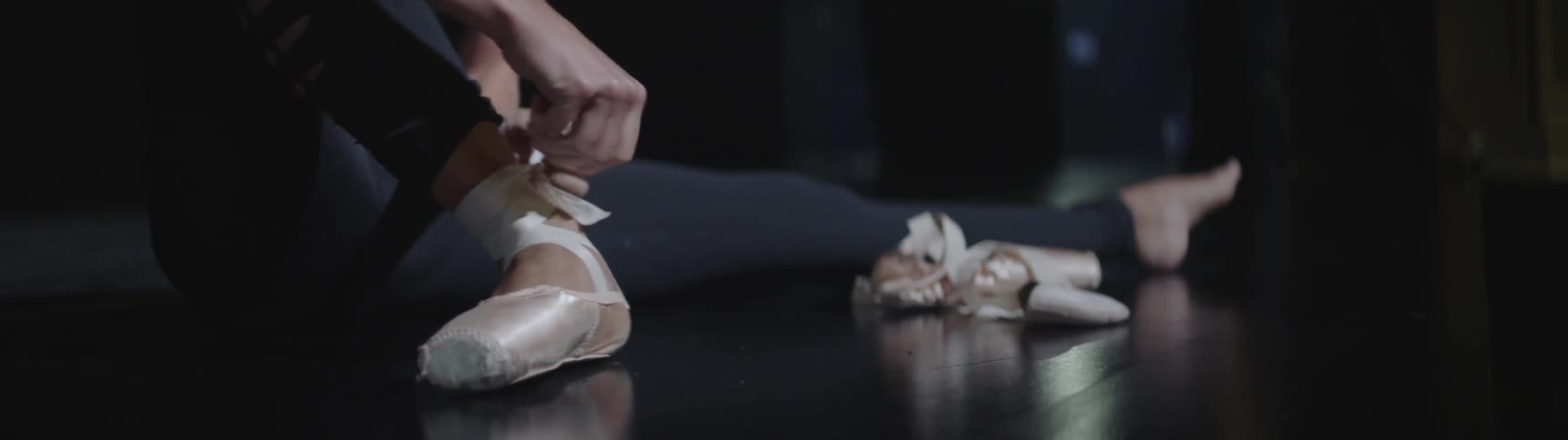 Ballet dancer tying ribbons on ballet shoes sitting on floor