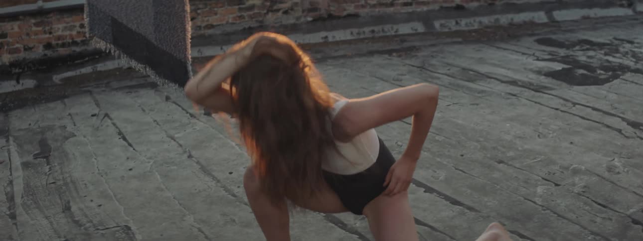 A female wearing a black and white leotard is practicing creative dance moves between two clothes lines on a rooftop while the sun is setting