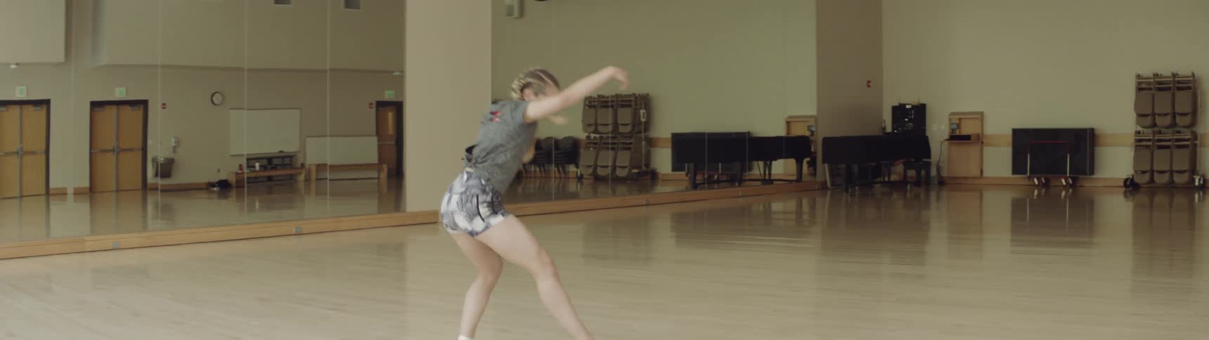 A young woman with blonde hair is dancing and raises one leg high in the air before turning taking a step and then doing a flip in a dance studio