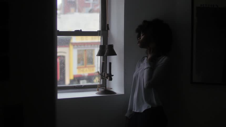 A woman stands at an apartment window looking outwards