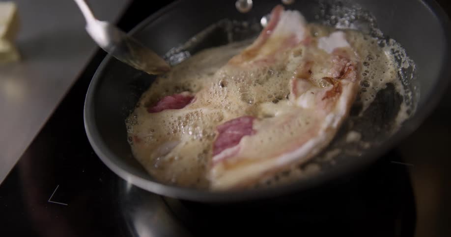 A person cooks bacon on a stove top, using a spoon to scoop up the hot, sizzling grease and pour it back onto the bacon in a slow motion and shaky close up shot.