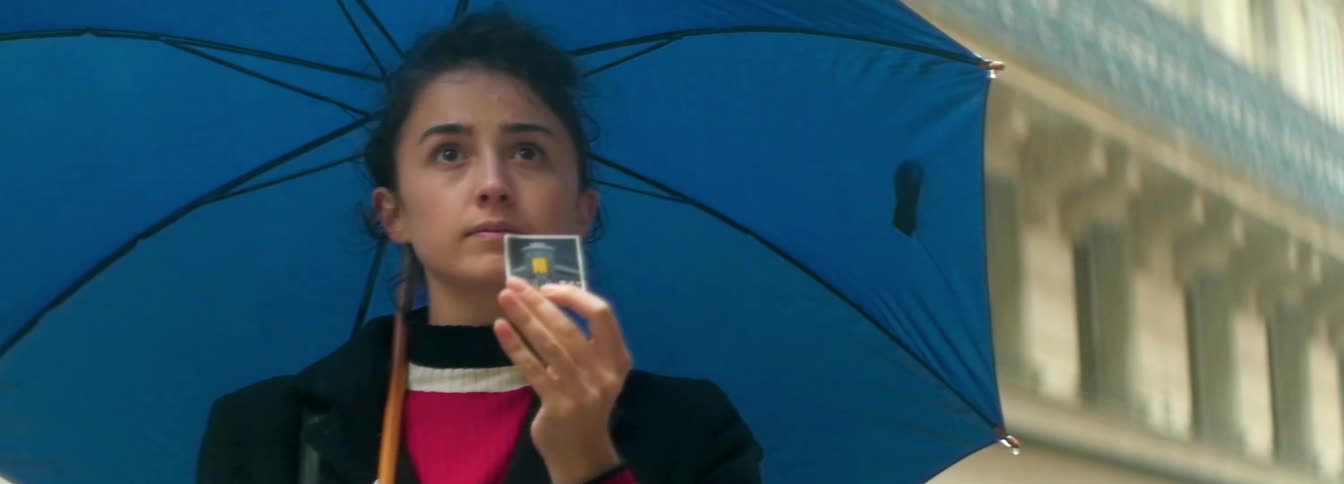 Woman carrying blue umbrella looks at card in front of a building in the city