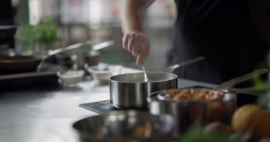 Bald cook wearing black apron stirs the cooking spaghetti in saucepan in the kitchen during daylight