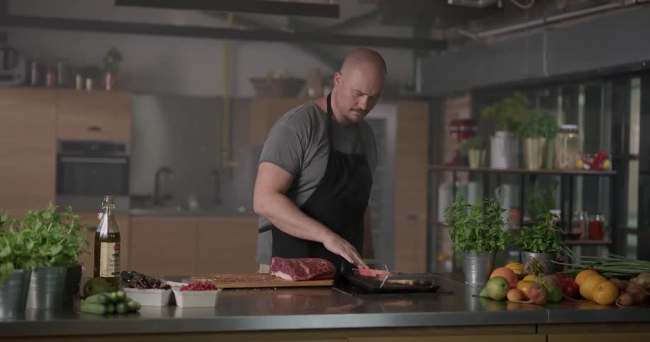 A white male seasons a piece of meat before putting it on a cooking flat top in a kitchen