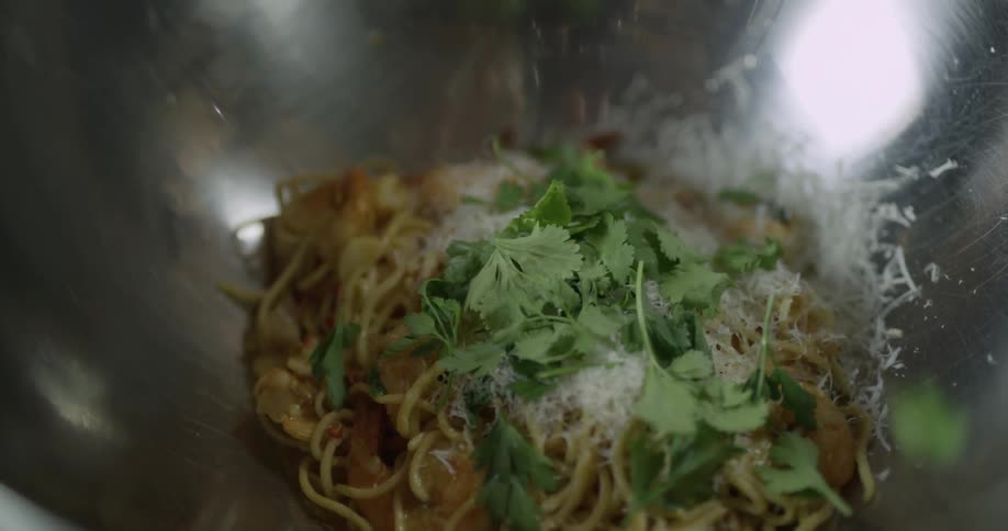Green herbs falling into a silver bowl of noodles
