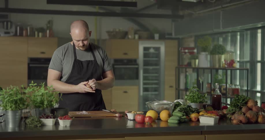 Bald headed cook wearing apron chops shallot on cutting board in the kitchen during daylight