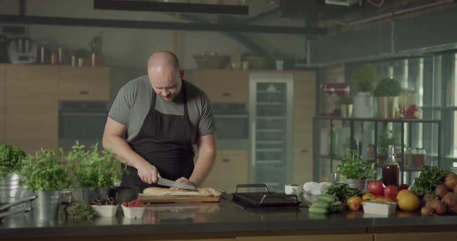 A white man slices a loaf of bread on a cutting board in a kitchen filled with planters of fresh herbs