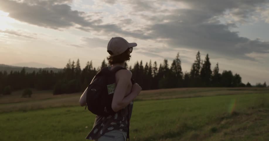 A white female hikes through a field with evergreens in the distance wearing a backpack hat and sunglasses