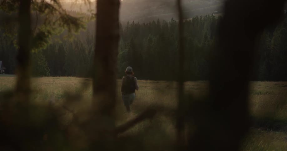 A woman in a hat walks out of a forest into a field at sunset