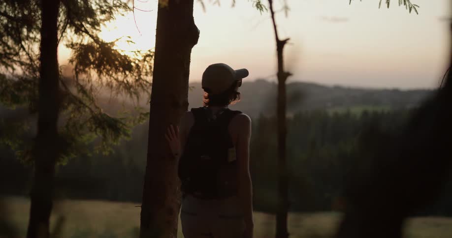 A woman in a hat walks out of a forest into a field at sunset