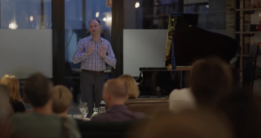 A man stands on a small stage before an audience of people and he talks aloud briefly before turning around to sit down at the grand piano behind him
