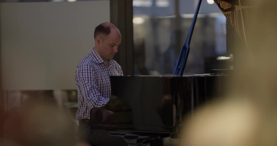 A white man in a plaid shirt plays a black baby grand piano