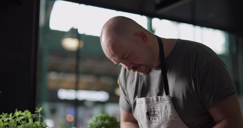 Bald headed man who wears white apron cuts fish on the cutting board in the kitchen during daylight