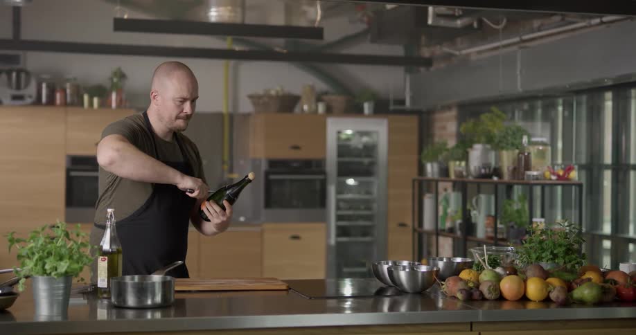 A bald man in a dark apron stands in a kitchen where he skillfully slides a knife up and down the neck of a bottle of champagne and actually manages to pop the cork without damaging the bottle and the champagne comes gushing out