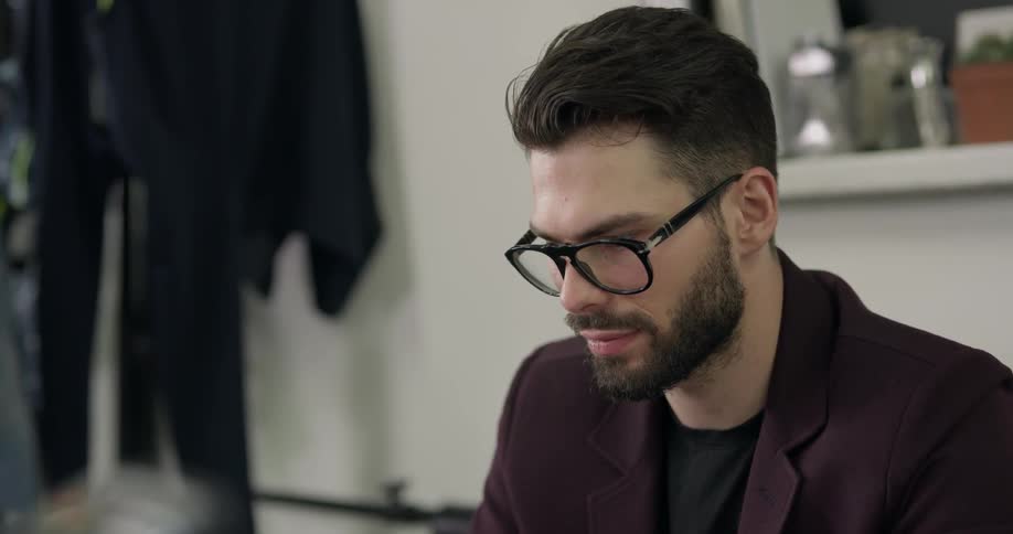 A young man with a beard and glasses is seated and concentrated closely on his task as he sits within a clothing store