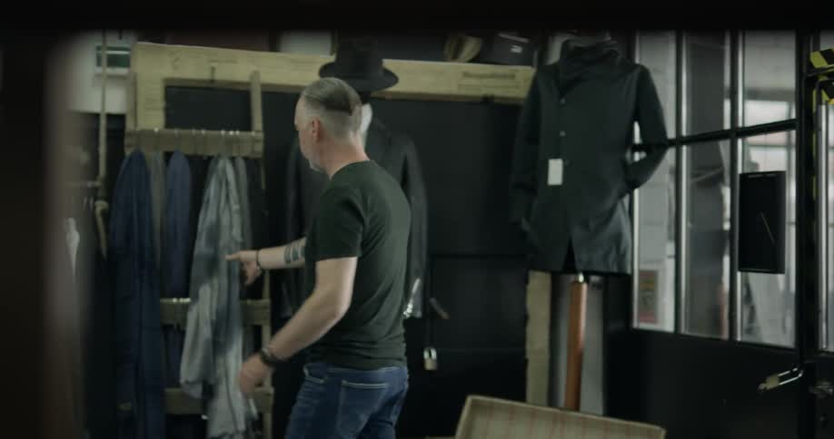 A white man with gray hair walks into a men's clothing store and begins browsing the clothing racks