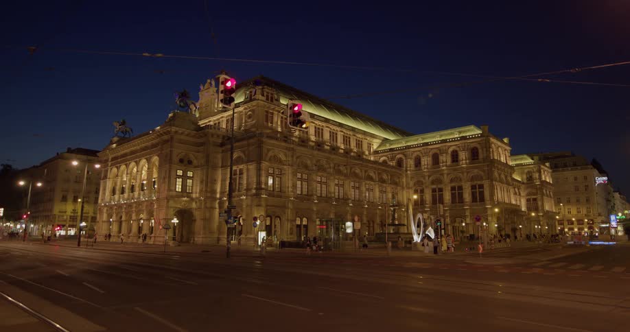 A large building with elaborate architecture stands before numerous pedestrians who are strolling nearby