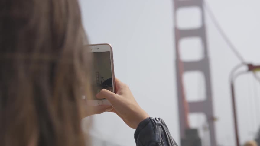 A woman holds up her phone and photographs the golden gate bridge on a windy day