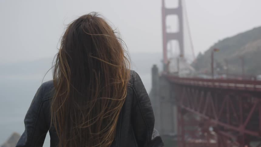 A brunette female has her hair blown in the wind next to the golden gate bridge