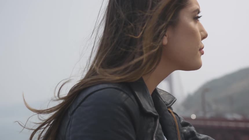 A woman stands near the golden gate bridge with her hair blowing in the wind