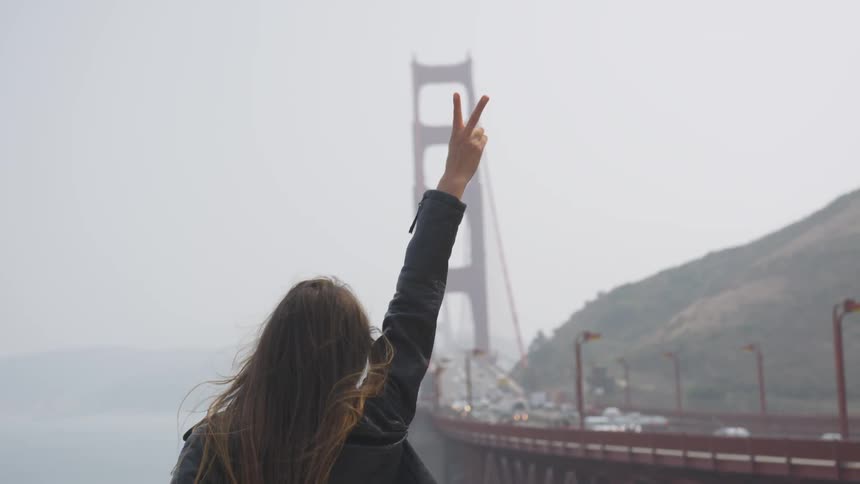 A woman flashes a peace sign while standing near the golden gate bridge on a foggy day