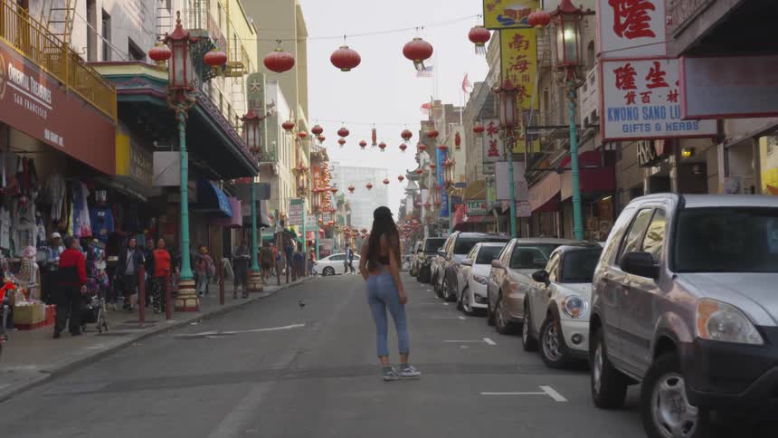 A woman walks down a city street and looks up and back at the hanging chinese lanterns and the asian signs on the buildings