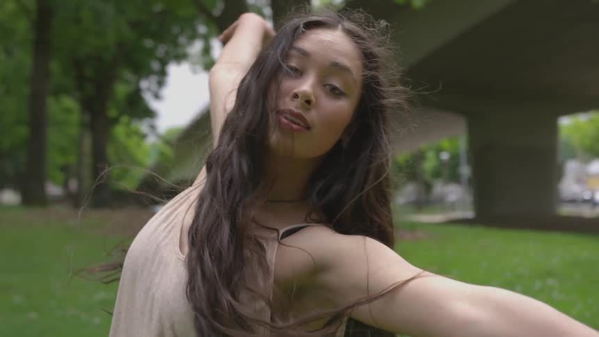 A brunette female dances in a sleeveless top on a lawn in an urban area