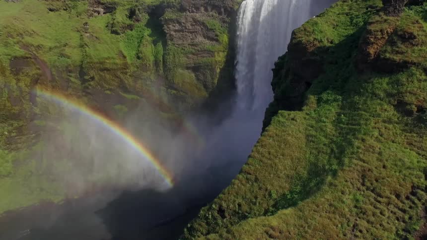 A rainbow forms out of w large waterfall against moss covered mountains