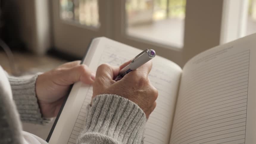 A female in a gray sweater is facing windows and writing in a journal with a pen