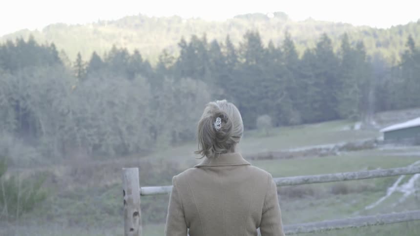 Blonde woman wearing cream coat walks down and takes look at the dense forest next to house in the daytime