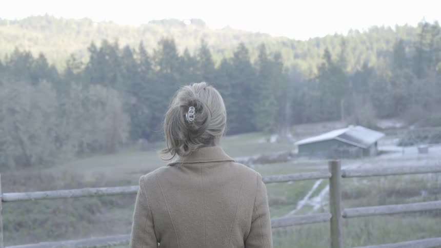 A woman stands next to a wood fence overlooking forest trees