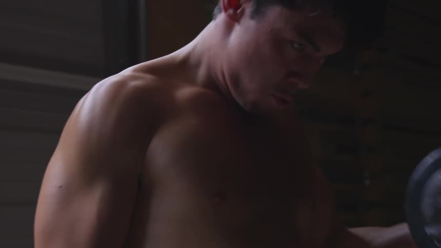 A shirtless man does alternating bicep curls in front of a garage door